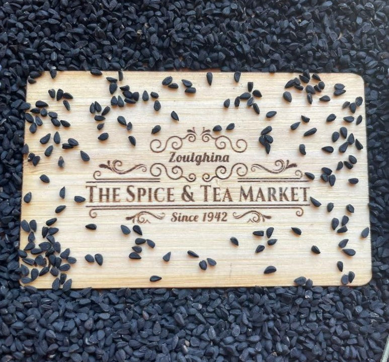 Wooden cutting board with nigella seeds and 'The Spice & Tea Market' branding on a dark textured background. حبة البركة