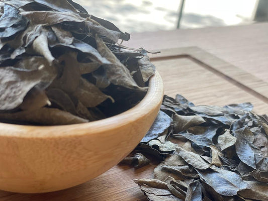 A wooden bowl filled with dried curry leaves on a wooden surface with some leaves scattered outside the bowl.